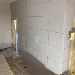 ART-DOM Tomasz Frankiewicz - Przygotowana ściana pod wykończenie, z widocznymi liniami poziomymi i pionowymi wyznaczonymi czerwoną kredką, zabezpieczone taśmą malarską narożniki i kable elektryczne, podłoga przykryta folią...