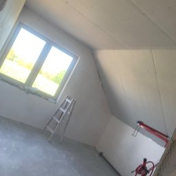 ART-DOM Tomasz Frankiewicz - Pokój na poddaszu w trakcie prac wykończeniowych, widoczne płyty gipsowo-kartonowe na skośnym suficie, okno dachowe, drabina i podnośnik do płyt.