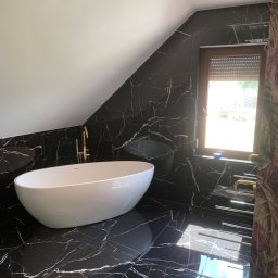 ART-DOM Tomasz Frankiewicz - Nowoczesna łazienka na poddaszu w czarnych marmurkowych płytkach z białą wolnostojącą wanną i złotą armaturą, oświetlona wpadającym światłem przez okno z roletami.