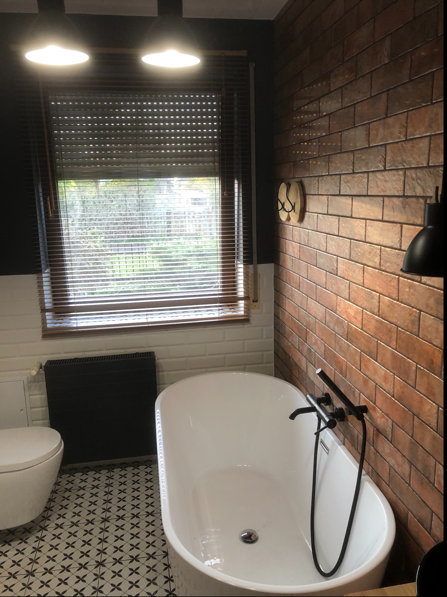 Nowoczesna łazienka z wolnostojącą białą wanną, czarną armaturą i grzejnikiem, ceglaną ścianą z dekoracyjnymi wieszakami, wzorzystą podłogą i oknem z żaluzjami.