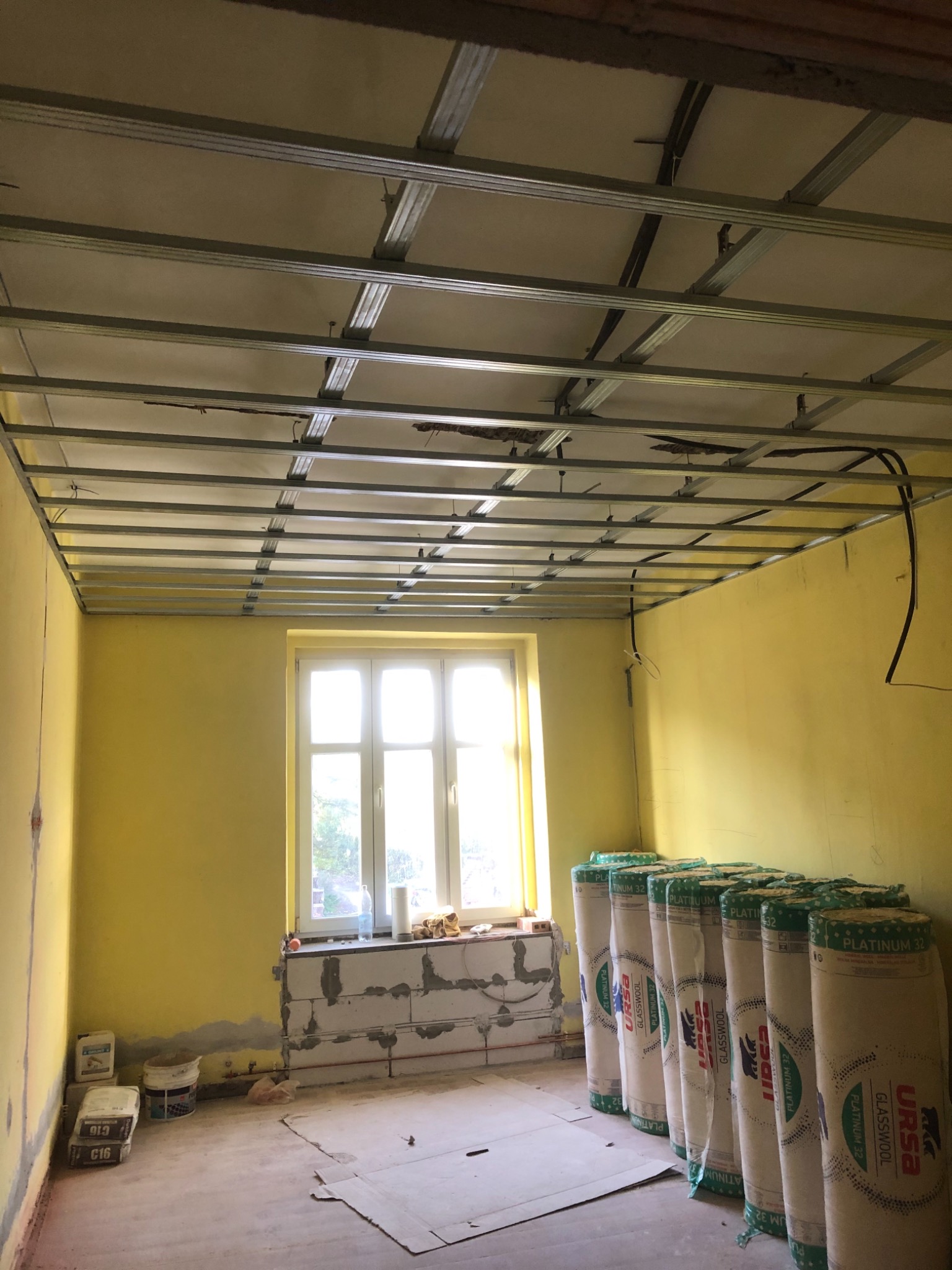 Metalowa konstrukcja sufitu podwieszanego w trakcie montażu, widoczne przewody elektryczne, żółte ściany, okno z parapetem z bloczków, rolki wełny mineralnej przy ścianie, przygotowania do prac...