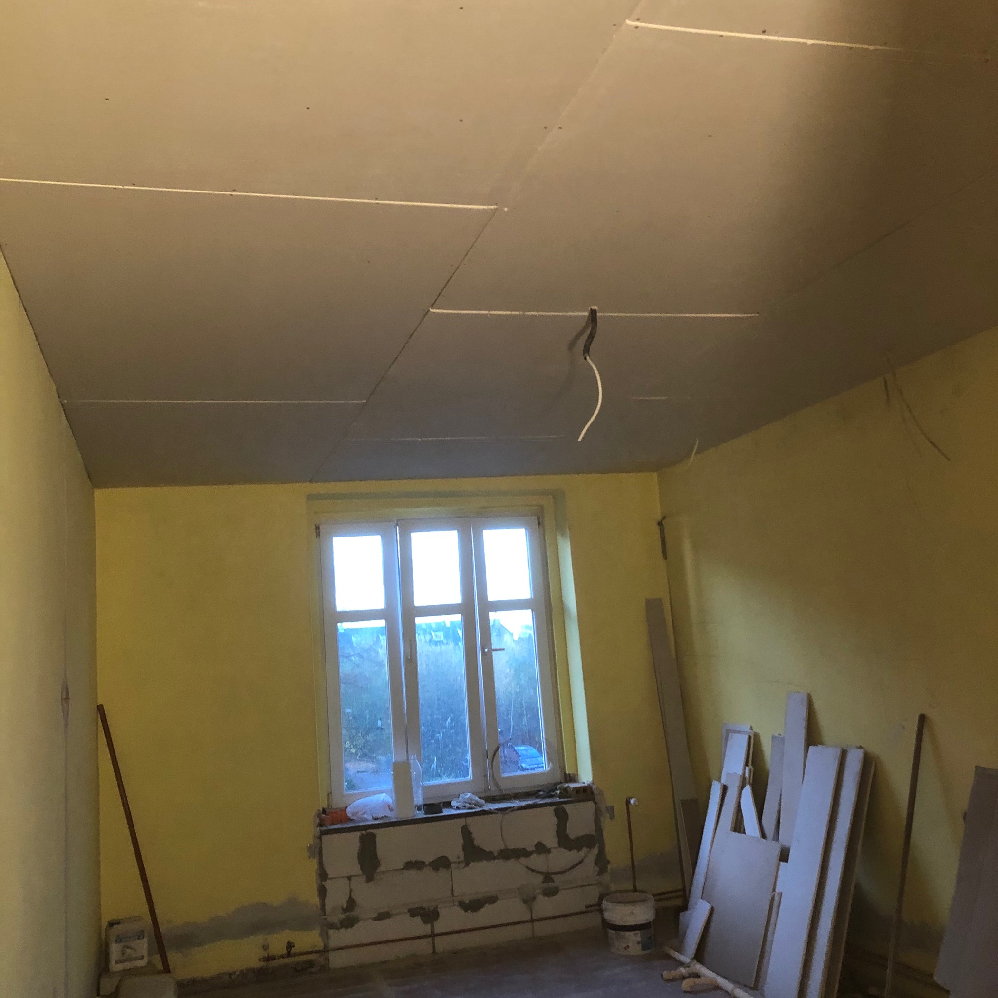 Wnętrze pokoju w trakcie remontu: żółte ściany, okno z widokiem na zewnątrz, częściowo zamontowany sufit z płyt kartonowo-gipsowych, widoczne przewody elektryczne, materiały budowlane przy ścianie.