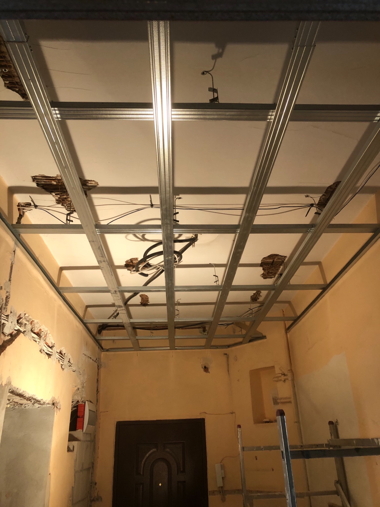 Montaż metalowej konstrukcji sufitu podwieszanego z widocznymi instalacjami elektrycznymi i uszkodzeniami starego sufitu, ściany w kolorze brzoskwiniowym, drabina w tle.