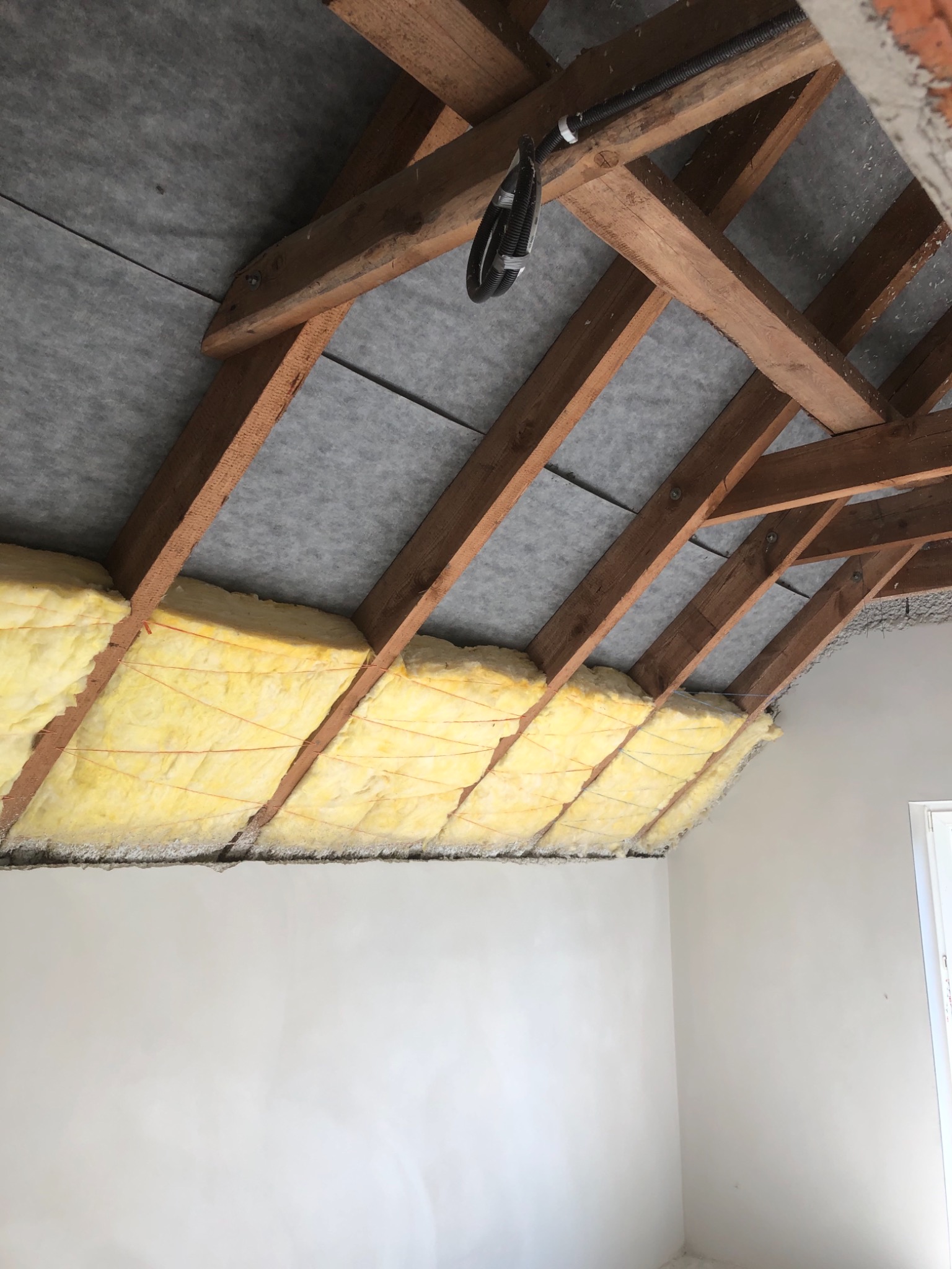 Konstrukcja drewniana poddasza z izolacją z wełny mineralnej, zabezpieczona szarą membraną, widoczne przewody elektryczne w peszlu.
