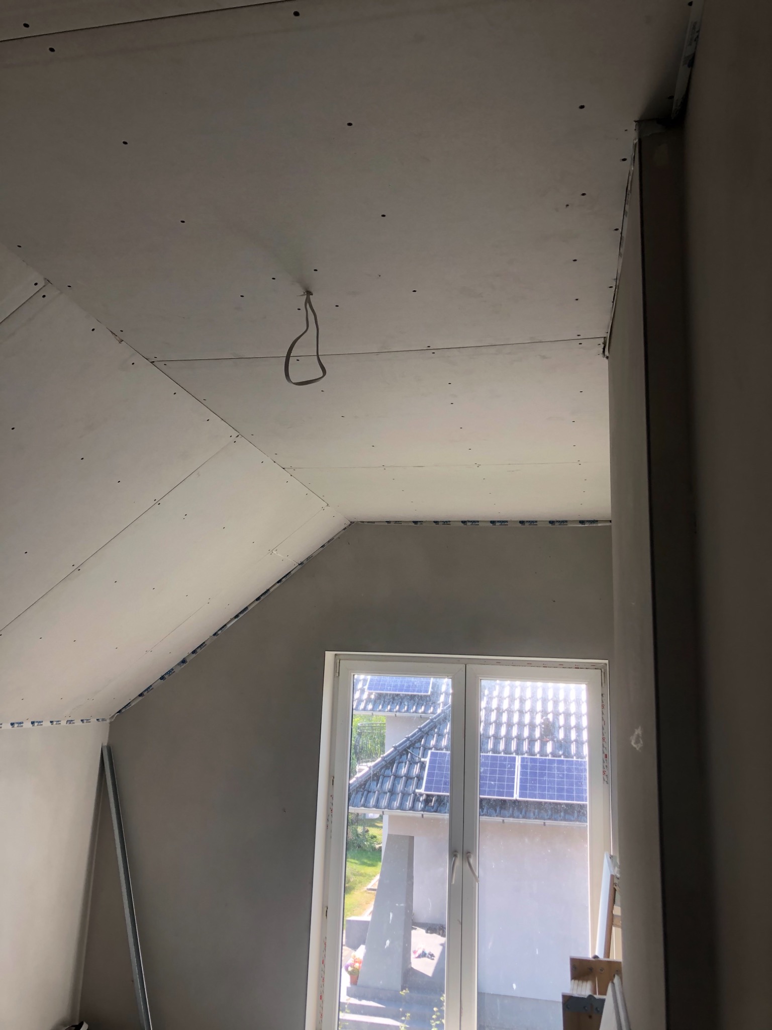 Poddasze w trakcie wykańczania płytami kartonowo-gipsowymi, widoczne łączenia płyt na skośnym suficie i ścianach, kabel zwisający z sufitu, okno z widokiem na dach z panelami słonecznymi.