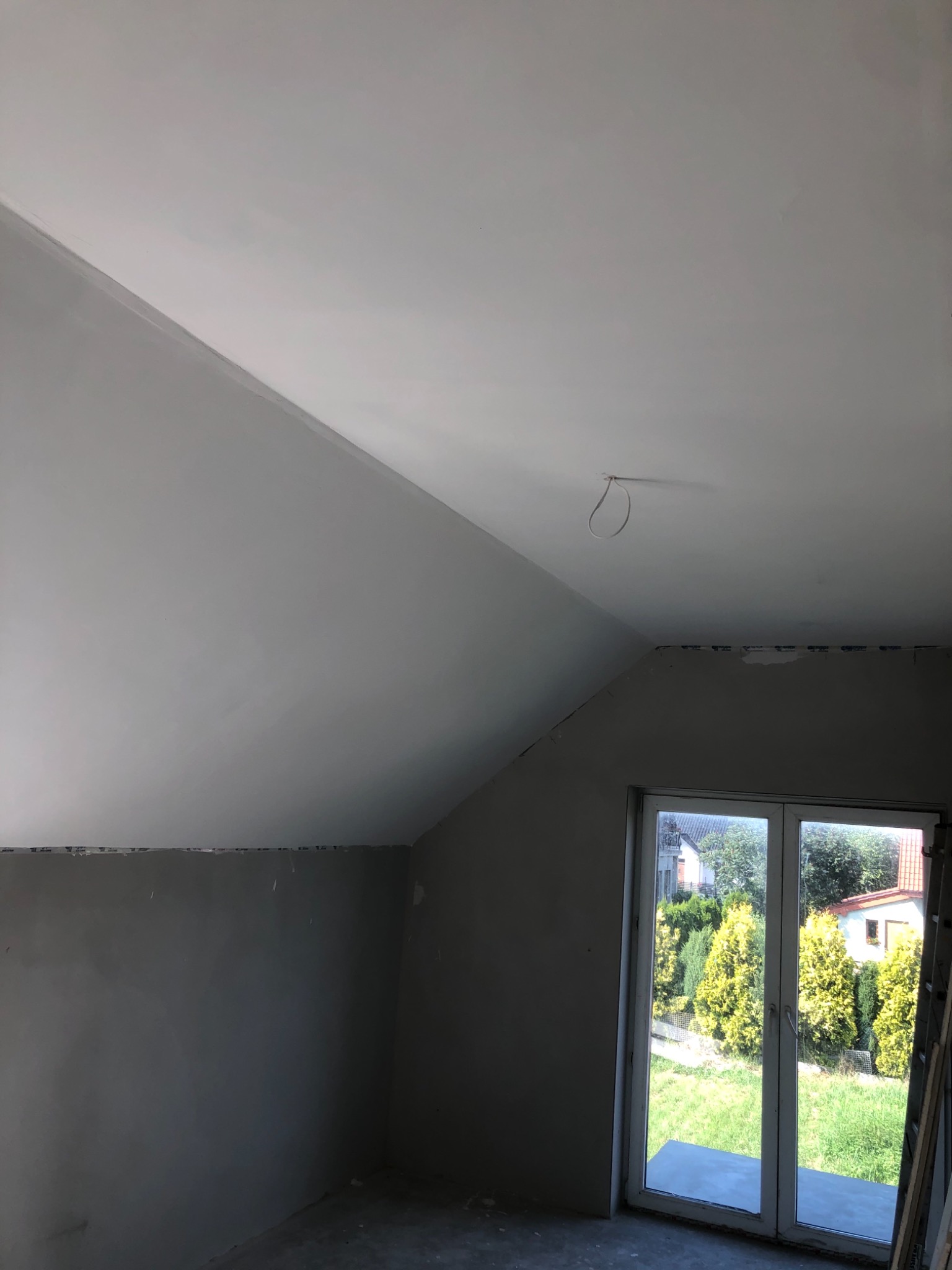 Pokój na poddaszu w trakcie szpachlowania ścian i sufitu, widoczne okno balkonowe z widokiem na ogród, surowy stan wykończenia.