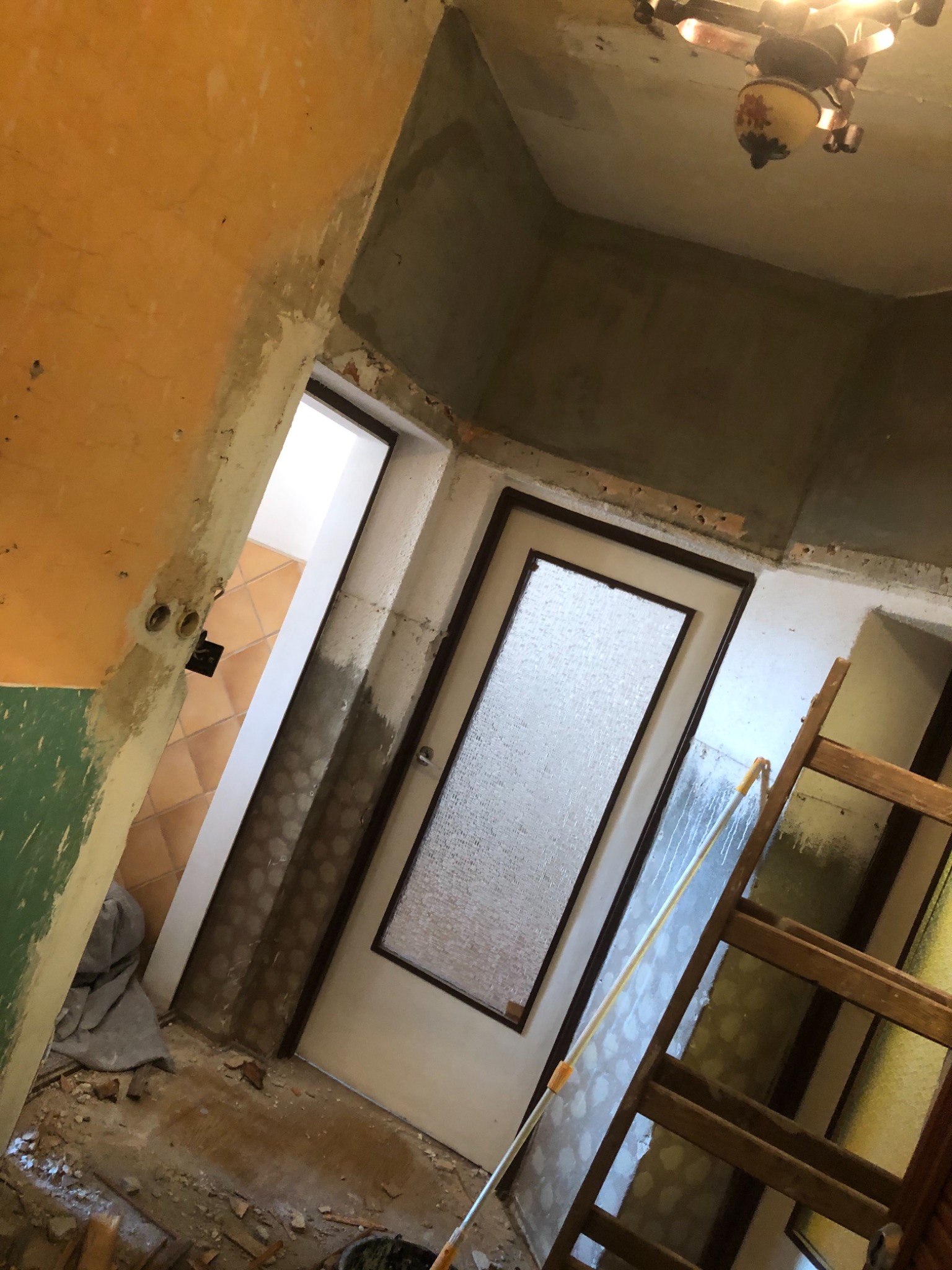 Wnętrze w trakcie remontu: stara futryna drzwiowa zmatowiona farbą, ściany z widocznymi śladami po skuciu tynku, stara drabina oparta o ścianę, widoczna instalacja elektryczna i fragment podłogi...