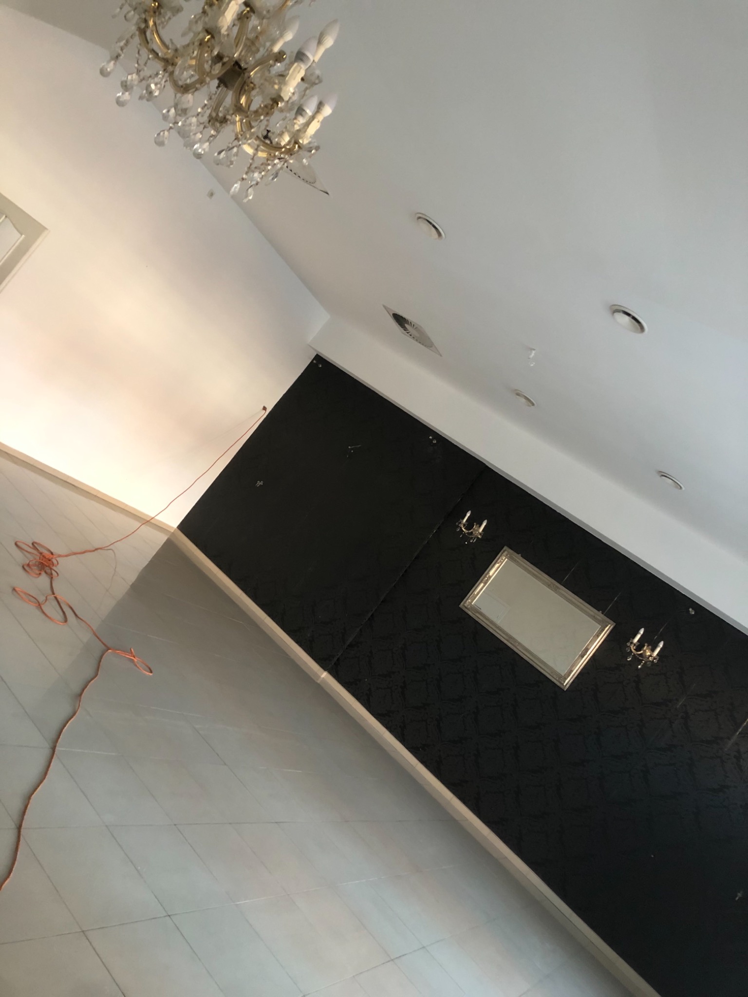 Wnętrze w trakcie remontu: czarna ściana z wzorem, lustro w złotej ramie, kinkiety, widoczna część sufitu z lampą i kabel elektryczny na podłodze z jasnych płytek.