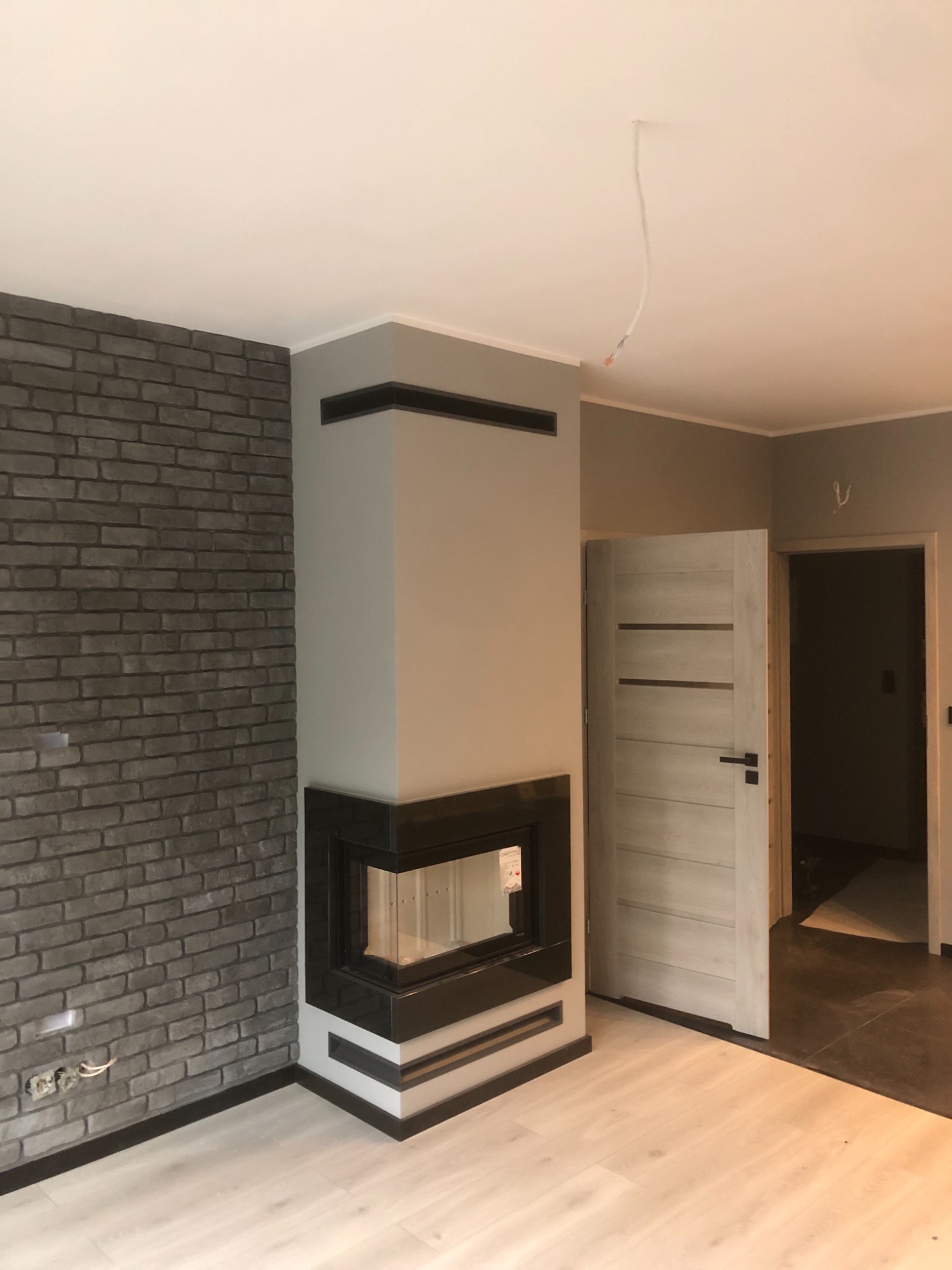 Wnętrze w Brzegu z kominkiem obudowanym szarą cegłą i czarnymi elementami, widoczne gniazdka elektryczne, biała podłoga i częściowo widoczne drzwi.
