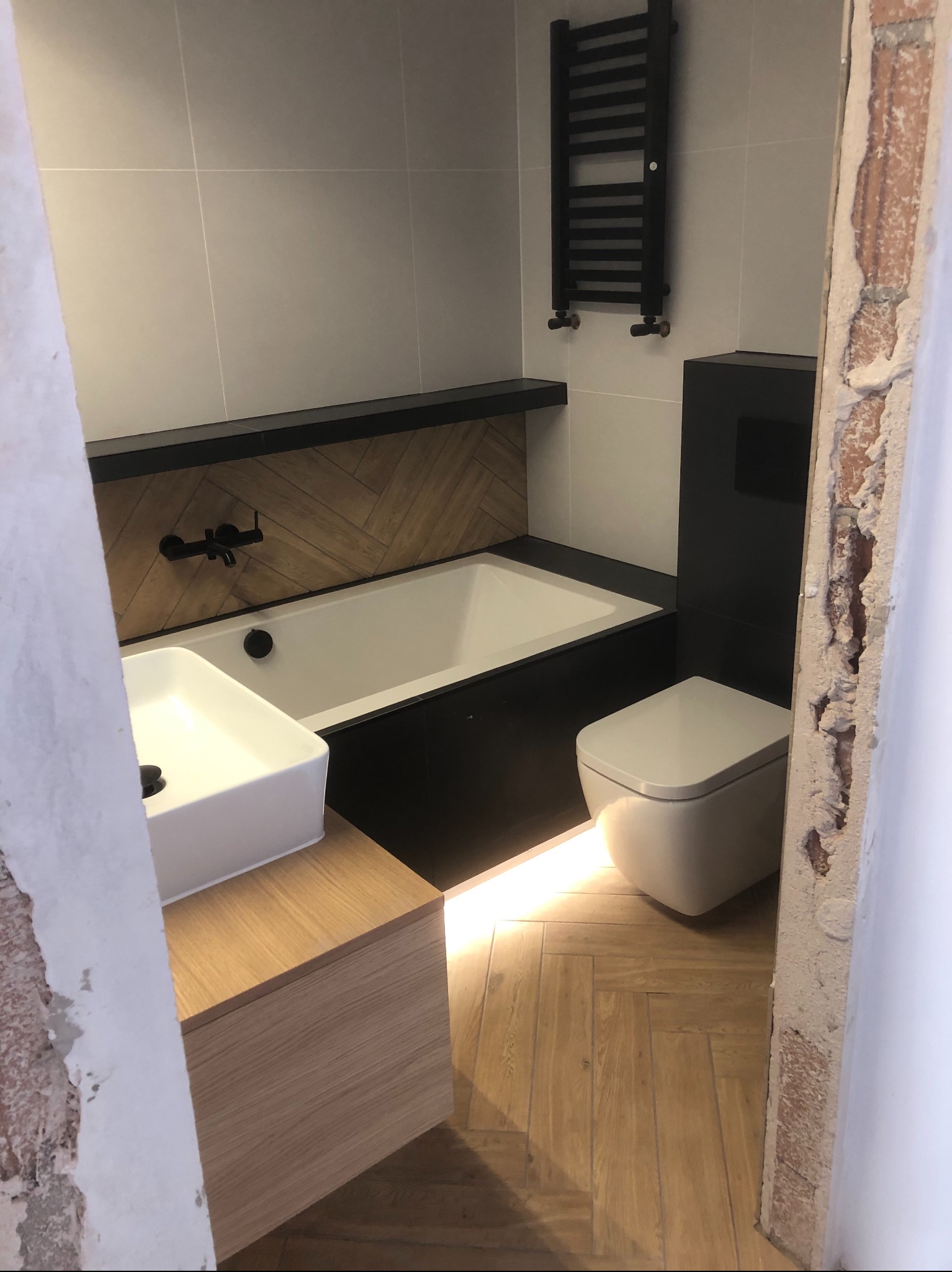 Nowoczesna łazienka w trakcie remontu, widoczna wanna z czarną obudową i drewnianym panelem dekoracyjnym, umywalka nablatowa na drewnianej szafce, toaleta oraz grzejnik drabinkowy na ścianie...