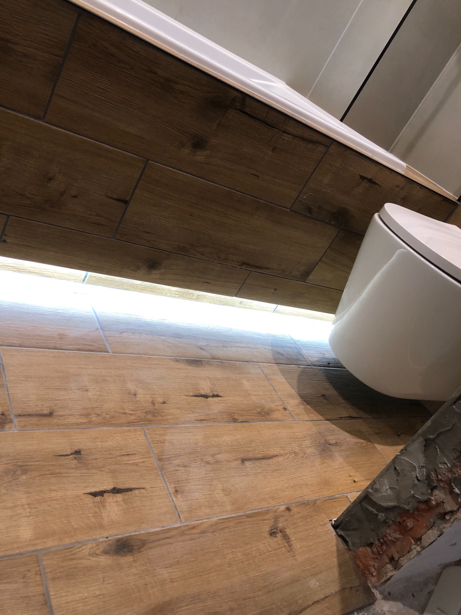 Ukończona łazienka z drewnopodobnymi płytkami na podłodze i ścianach, podwieszana toaleta, listwa LED pod wanną i fragmentem surowej ściany z cegły.