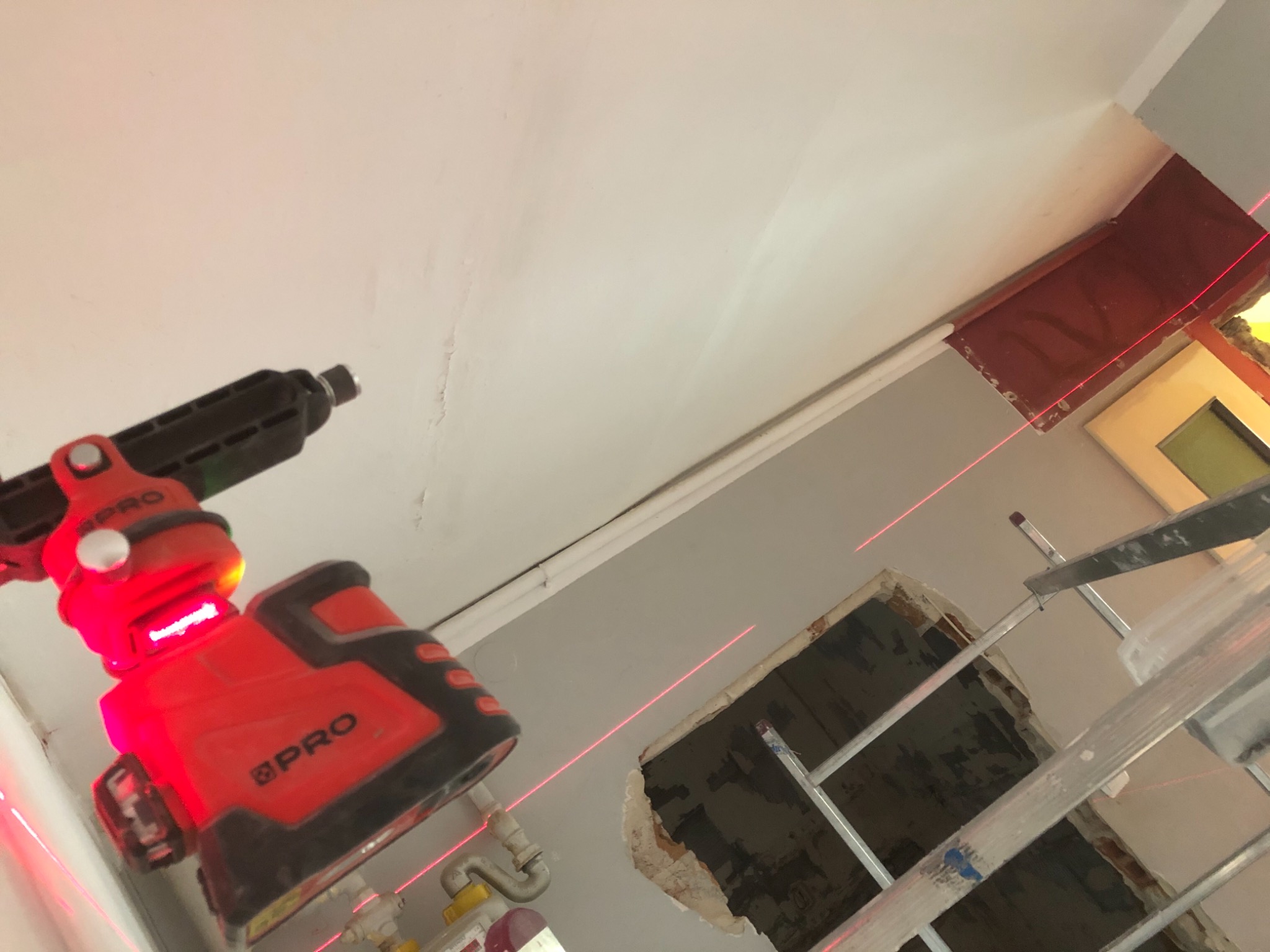 Czerwony laser krzyżowy wyznacza poziom na suficie podczas remontu, widoczna drabina i fragment ściany z otworem.