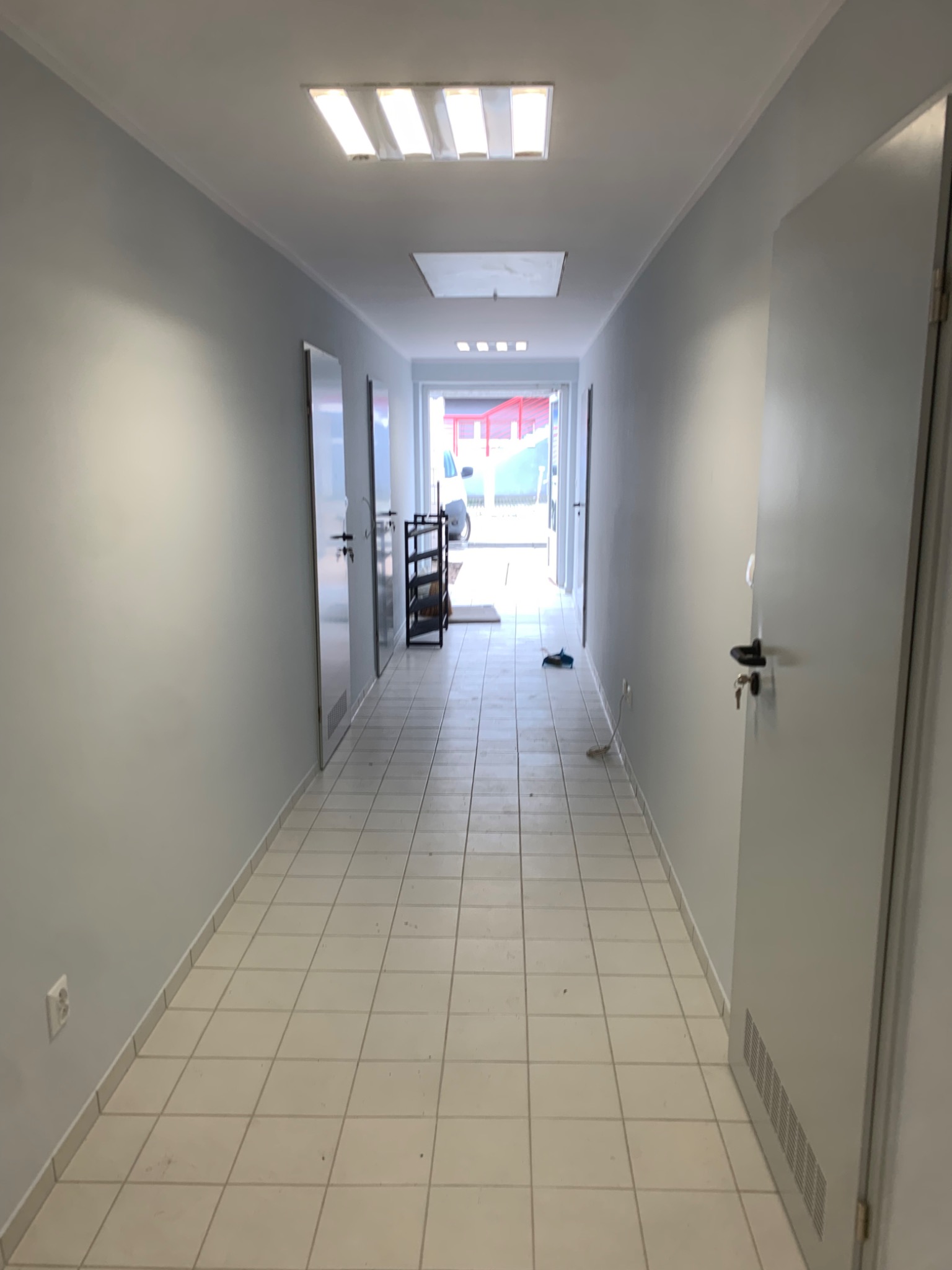 Długi, jasny korytarz biurowy z białymi ścianami i podłogą z kwadratowych płytek. Widoczne drzwi po obu stronach, oświetlenie sufitowe i regał na buty w oddali.