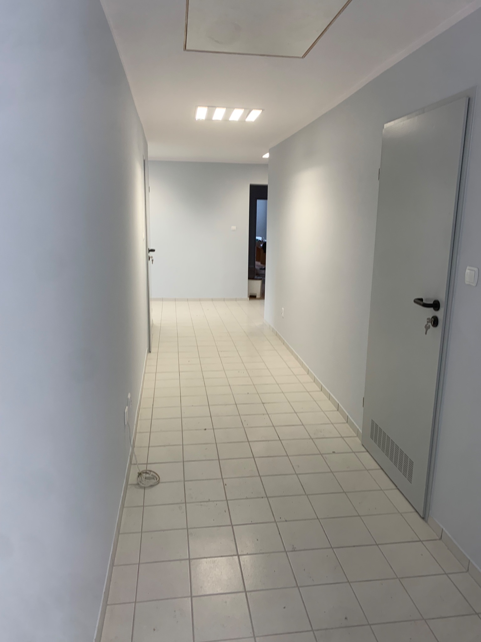 Korytarz biurowy po remoncie: świeżo pomalowane ściany na szaro, biała podłoga z płytek, białe drzwi z czarnymi klamkami i kluczem w zamku, sufit z wbudowanym oświetleniem