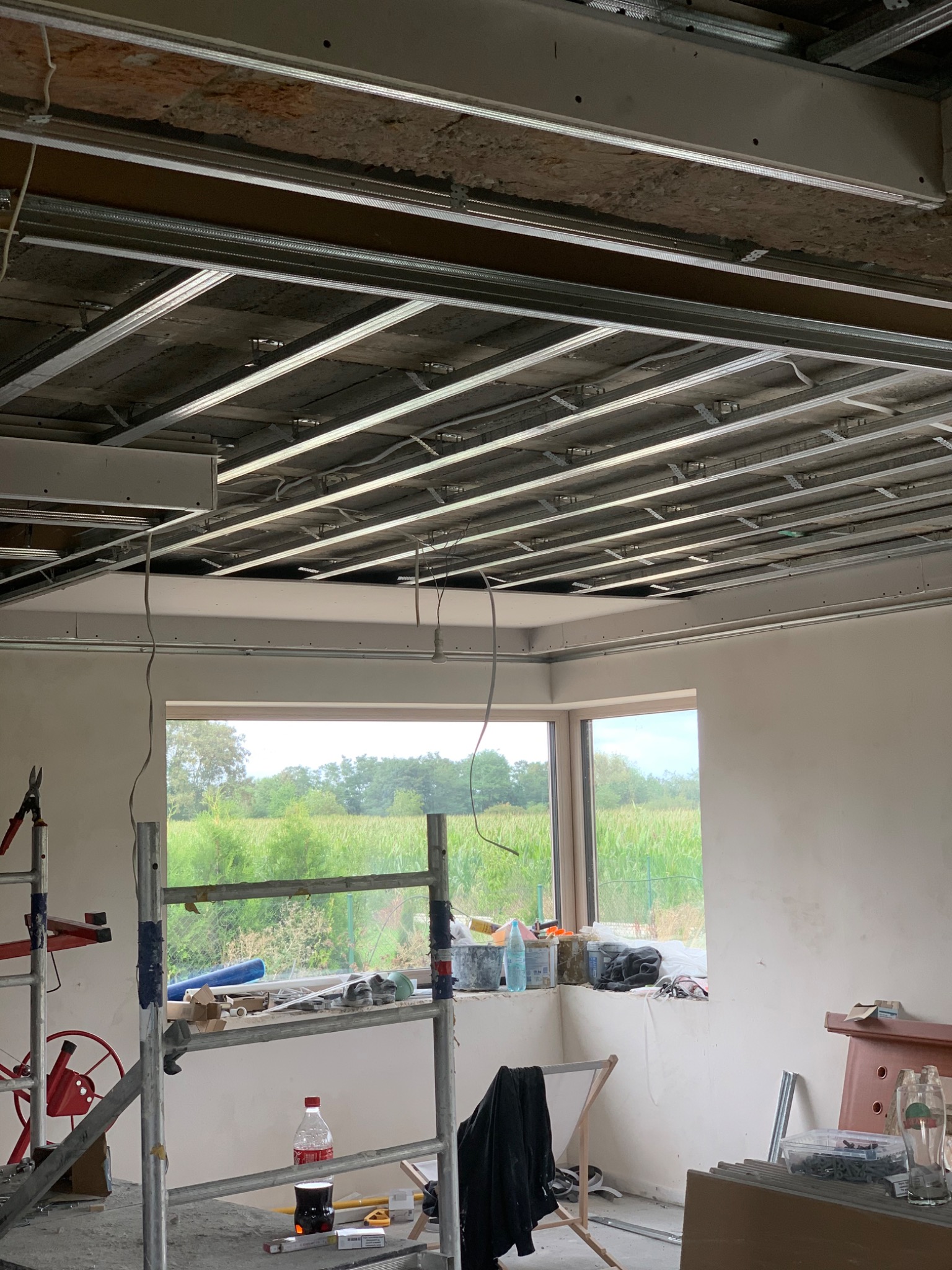 Montaż metalowego stelażu sufitu podwieszanego w pomieszczeniu z widokiem na zielone pole przez okno, widoczne narzędzia i materiały budowlane na rusztowaniu.