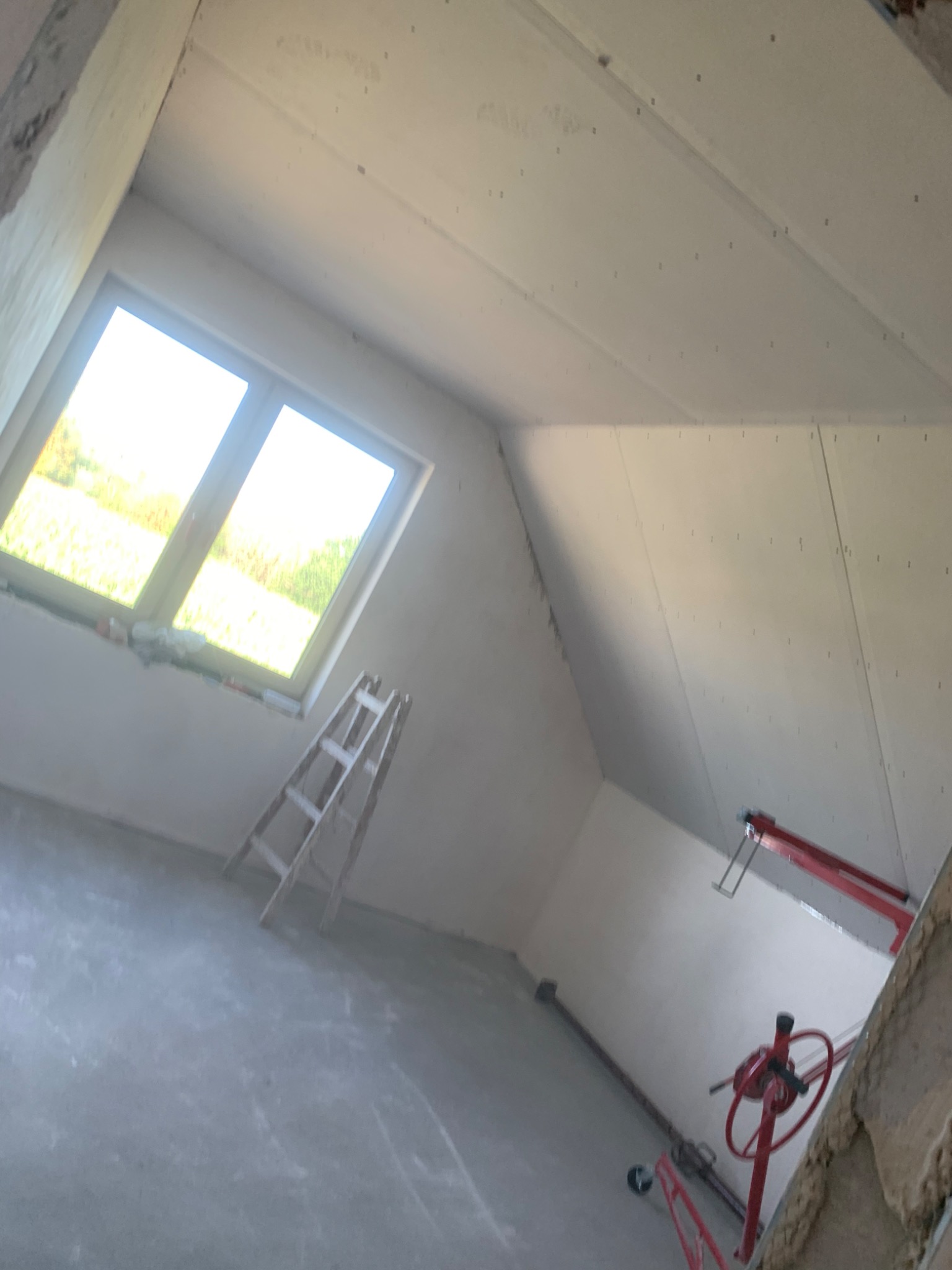 Pokój na poddaszu w trakcie prac wykończeniowych, widoczne płyty gipsowo-kartonowe na skośnym suficie, okno dachowe, drabina i podnośnik do płyt.