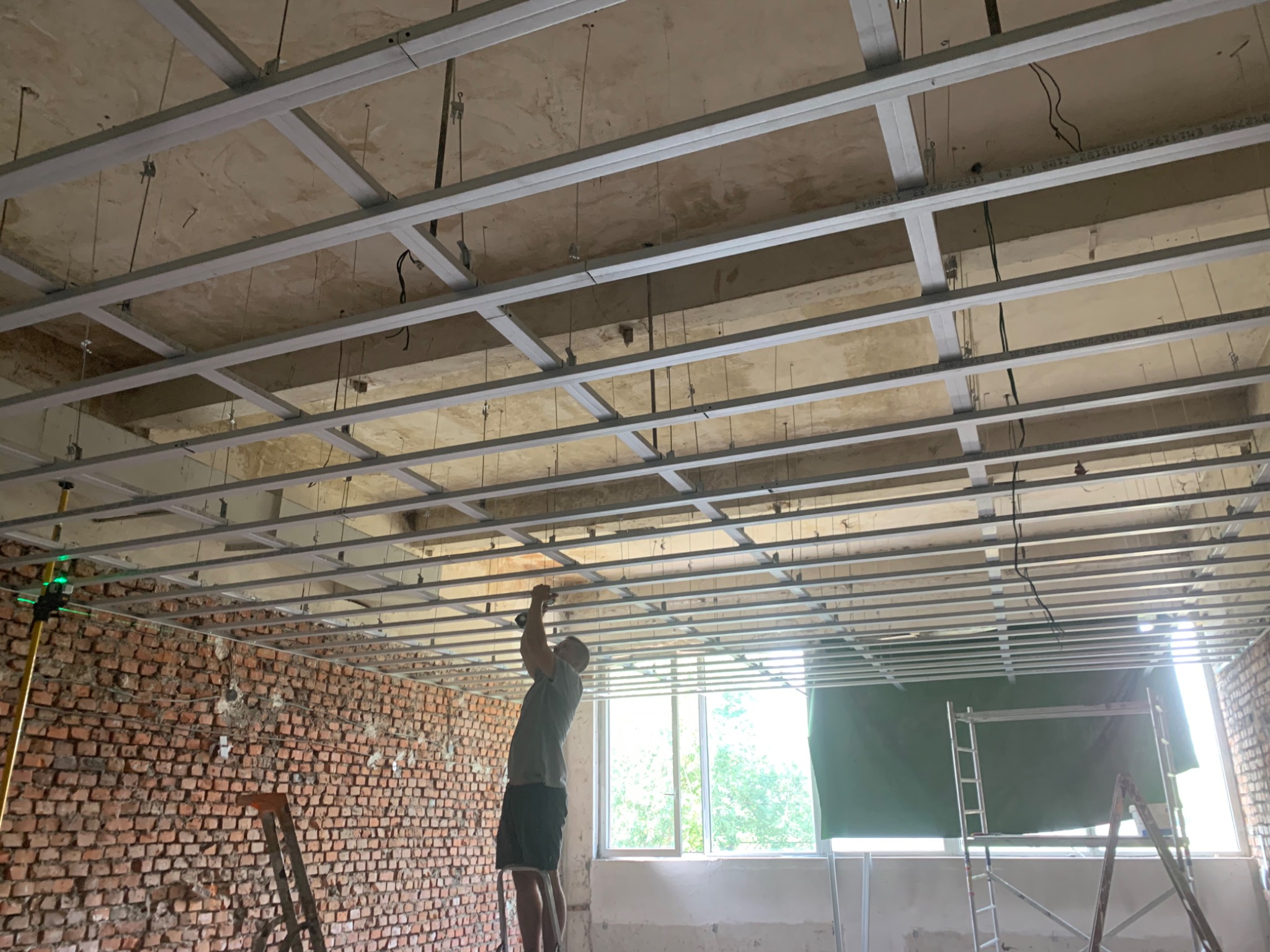 Montaż metalowej konstrukcji sufitu podwieszanego w surowym wnętrzu z odsłoniętą cegłą i laserowym niwelatorem.