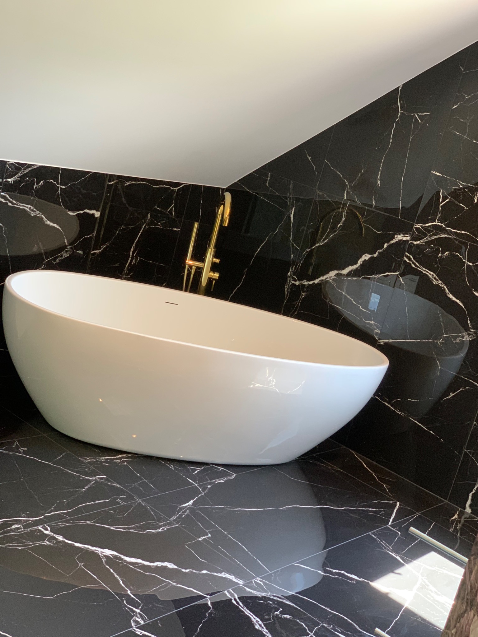 Nowoczesna, owalna wanna wolnostojąca z złotą armaturą na tle ściany i podłogi wyłożonej czarnym marmurem z białym żyłkowaniem, widoczne odbicie światła na powierzchni płytek