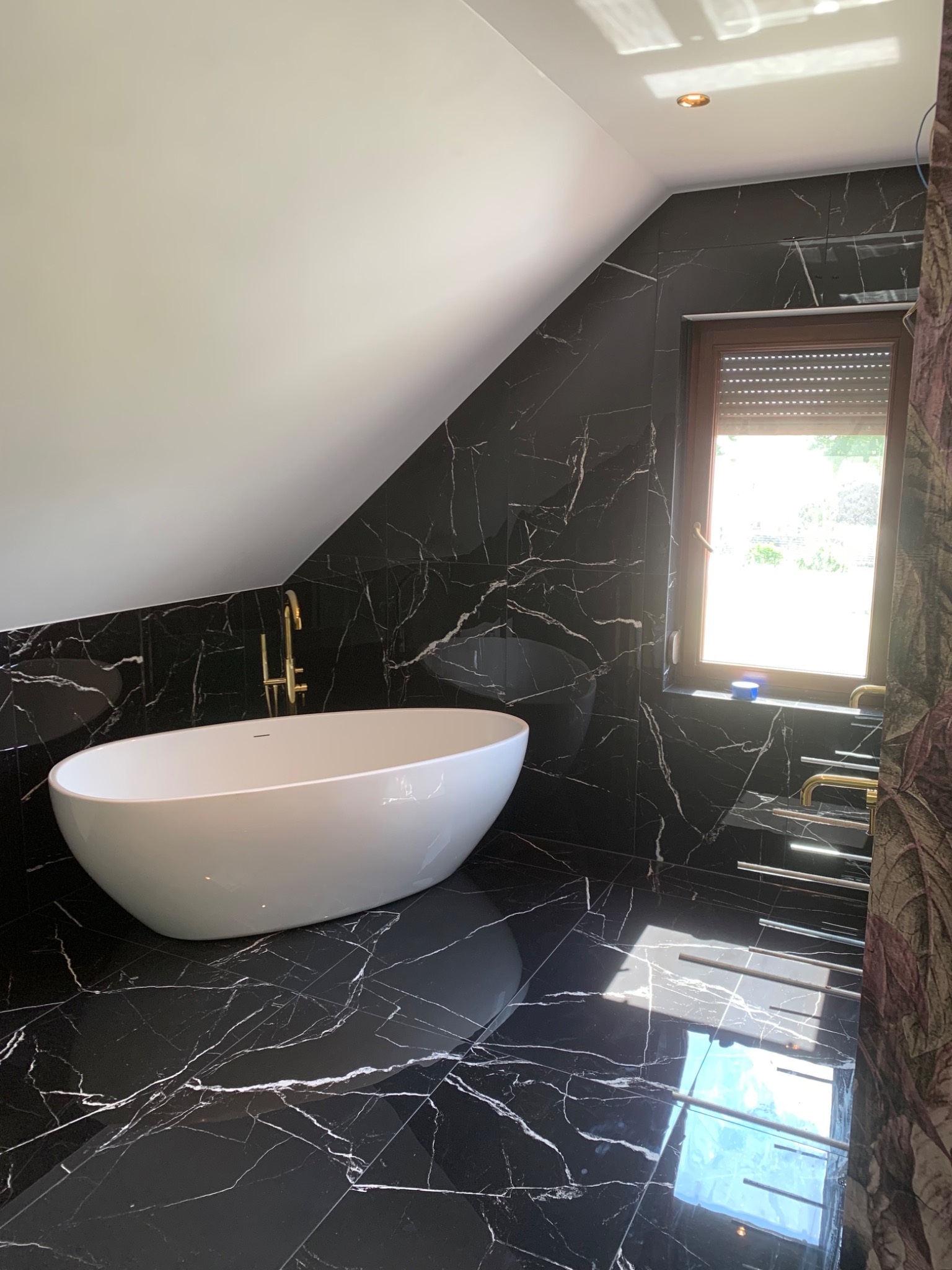 Nowoczesna łazienka na poddaszu w czarnych marmurkowych płytkach z białą wolnostojącą wanną i złotą armaturą, oświetlona wpadającym światłem przez okno z roletami.