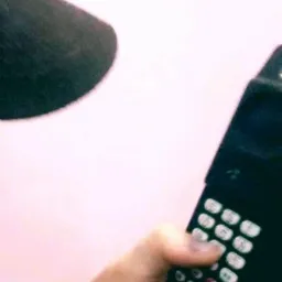 Dłoń naciska przyciski na czarnym telefonie komórkowym z anteną, widoczny fragment mikrofonu i różowe tło.