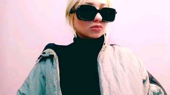 Osoba w czarnym golfie i oversize'owej kurtce, z dużymi, czarnymi okularami przeciwsłonecznymi, na tle różowej ściany.