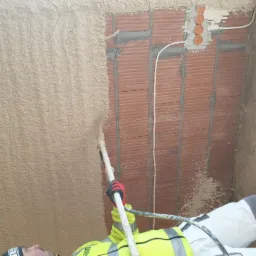 Aplikacja tynku maszynowego na ścianę z cegły, widoczny fragment instalacji elektrycznej i pracownik w odzieży roboczej, w trakcie pracy z użyciem specjalistycznego sprzętu.