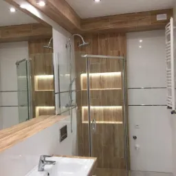 Nowoczesna łazienka z kabiną prysznicową typu walk-in, umywalką nablatową z drewnopodobnym blatem, lustrem i podświetlonymi półkami wewnątrz kabiny.