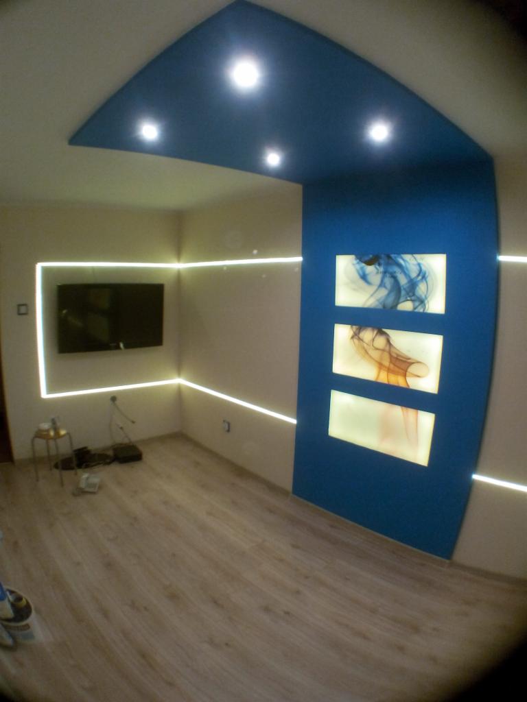 Nowoczesny pokój z podświetlanymi panelami ściennymi, niebieskim sufitem podwieszanym z oświetleniem punktowym i telewizorem, oświetlony listwami LED przy suficie i podłodze.