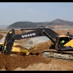 Żółto-czarny koparka Volvo EC750EL ładuje ziemię do żółtej wywrotki przegubowej Volvo na placu budowy z pagórkami i budynkami w tle.