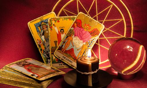 Rozłożone karty tarota, płonąca świeca na czarnym talerzyku, kryształowa kula i złoty ornament na tle bordowej tkaniny.