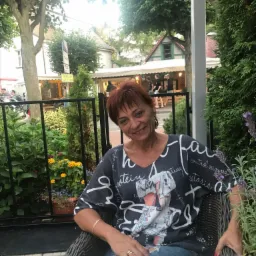 Uśmiechnięta kobieta z krótkimi rudymi włosami siedzi w wiklinowym fotelu na zewnątrz, na tle zieleni i budynku z szyldem Taxi 24h.