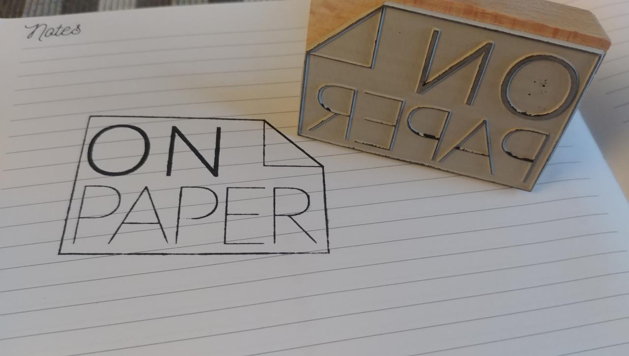 Stempel z napisem 'ON PAPER' odbity na liniowanym papierze notatnika. Obok leży stempel z odwróconym napisem.
