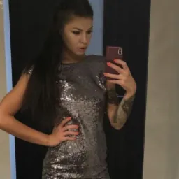 Kobieta robi selfie w lustrze, ubrana w srebrną, cekinową sukienkę mini, z tatuażem na ramieniu i pomalowanymi na pomarańczowo paznokciami.