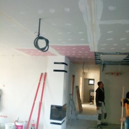 R. J - Wnętrze w trakcie remontu: widoczne płyty kartonowo-gipsowe na suficie, instalacja elektryczna zwisająca z sufitu, poziomice oparte o ścianę, biały kominek z czarnymi akcentami, pracownik...