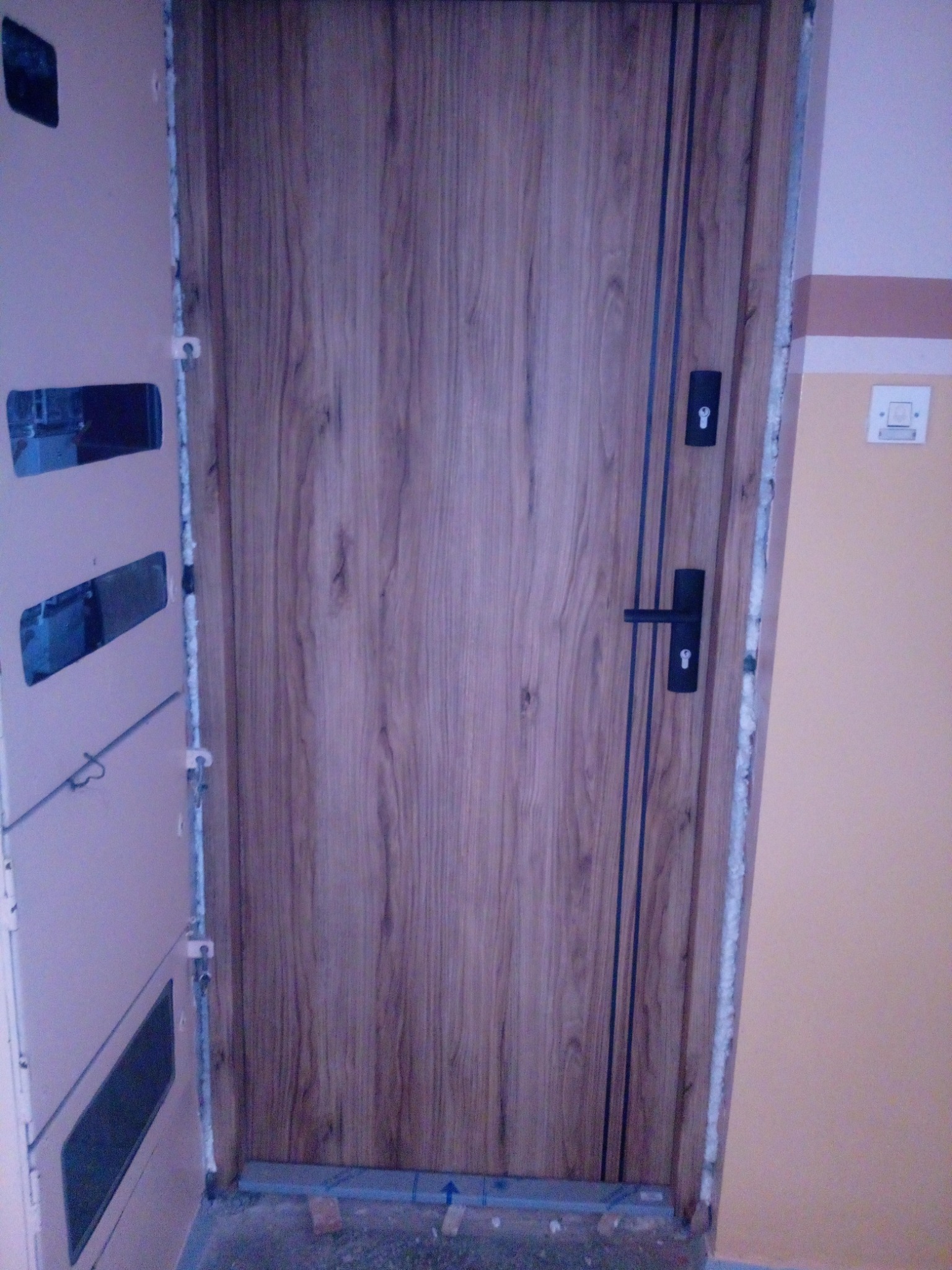 Montaż nowych drzwi wewnętrznych z drewnopodobnym wykończeniem i czarnymi akcentami, widoczne pianka montażowa i fragment ściany z płyt kartonowo-gipsowych.