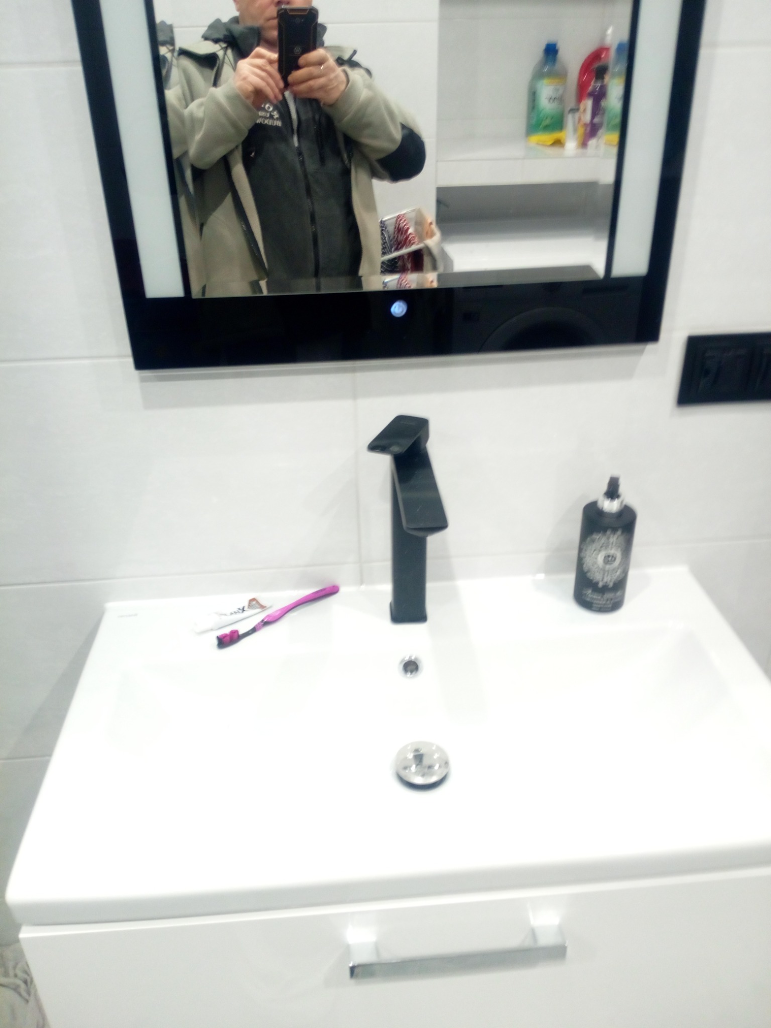 Biała umywalka z czarną baterią i akcesoriami w nowoczesnej łazience z białymi płytkami, odbicie fotografa w lustrze.