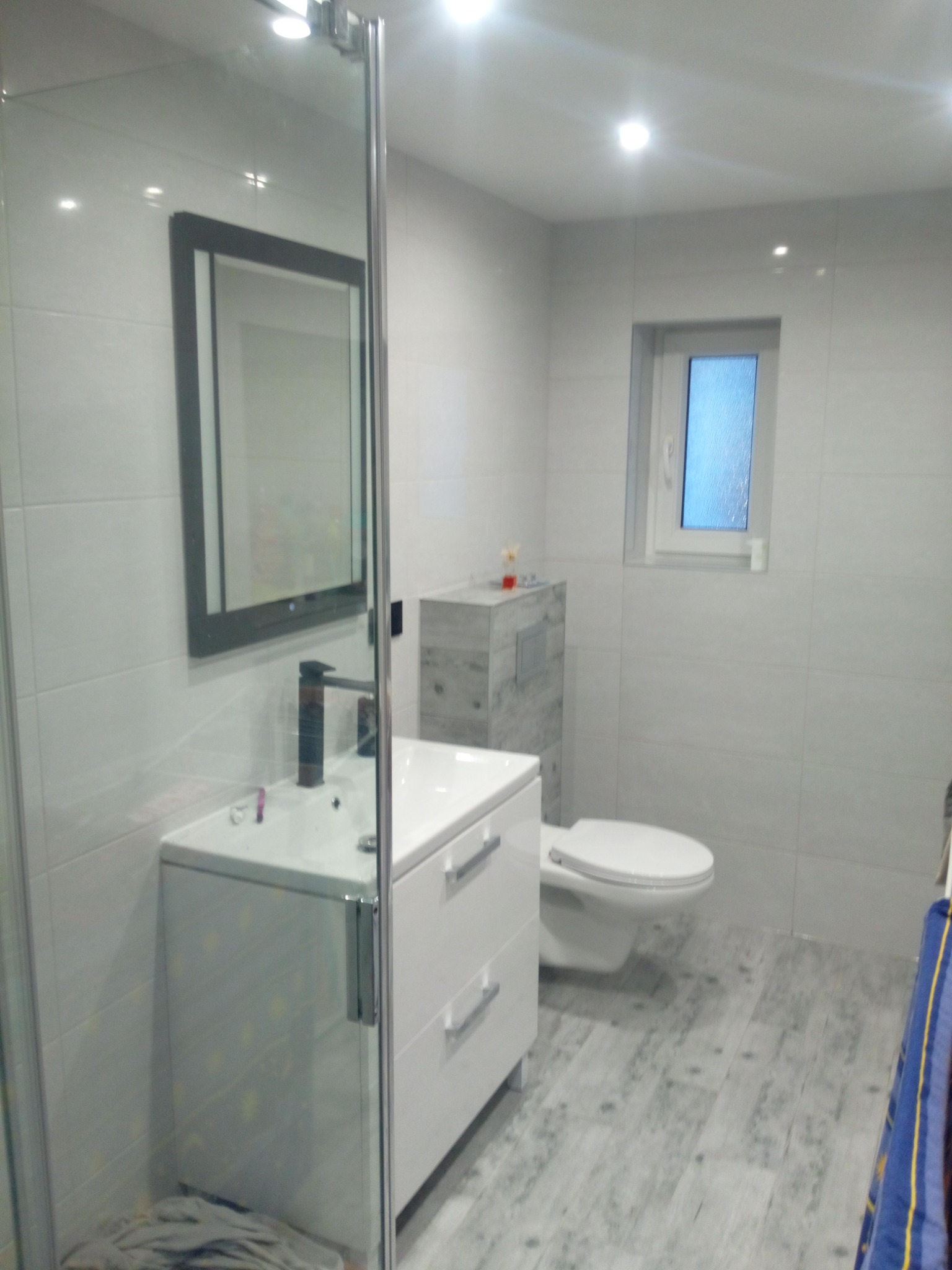 Jasna łazienka z kabiną prysznicową, umywalką z szafką, toaletą i oknem, wykończona płytkami w odcieniach szarości, z podłogą imitującą drewno.