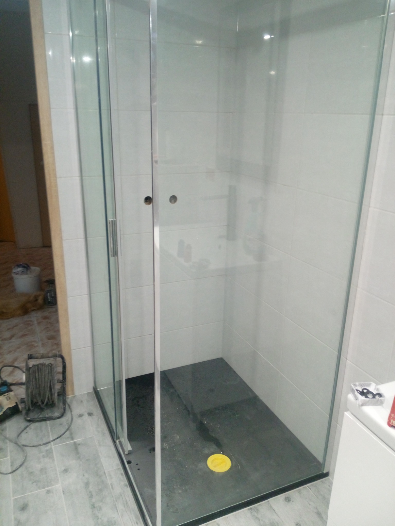 Kabina prysznicowa z czarnym brodzikiem i przezroczystymi szklanymi ściankami, widoczna na tle białych płytek ściennych i szarych płytek podłogowych, z widocznymi elementami instalacyjnymi...