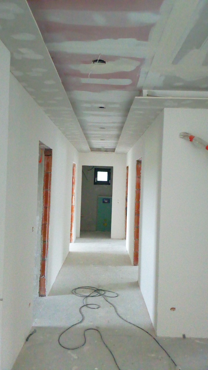 Korytarz w trakcie remontu z widoczną konstrukcją sufitu podwieszanego z płyt kartonowo-gipsowych, ściany przygotowane do malowania, widoczne kable na podłodze.