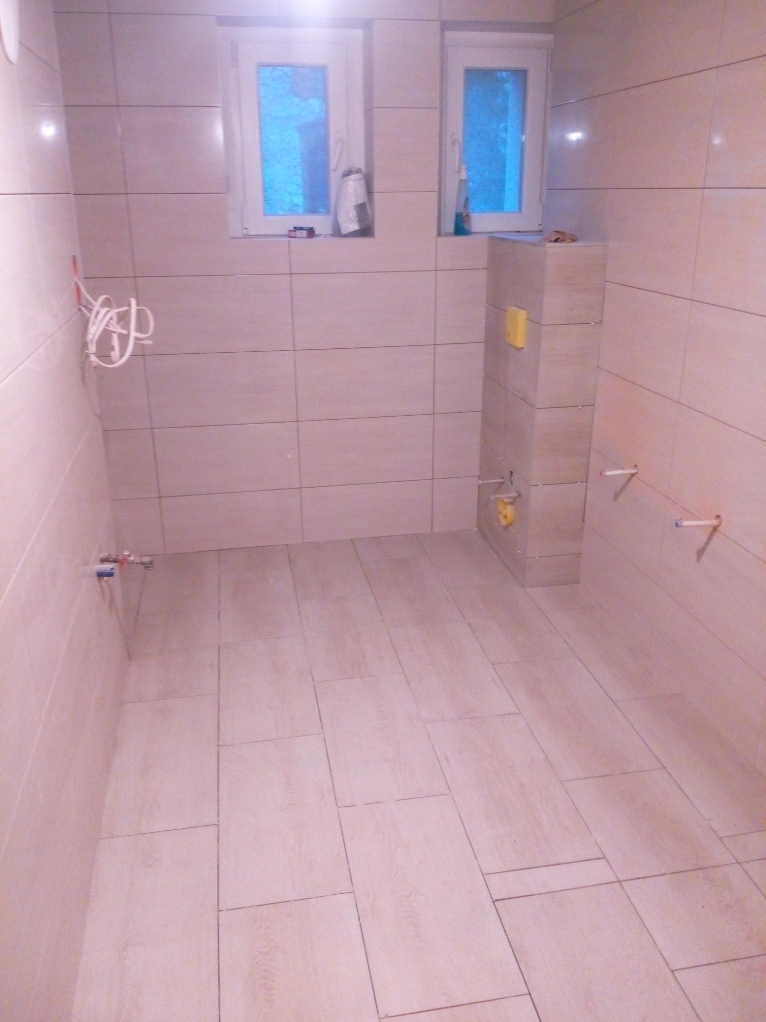 Wnętrze łazienki w Jemielnicy w trakcie prac glazurniczych, widoczne ułożone beżowe płytki na ścianach i podłodze, przygotowane instalacje wodne i sanitarna.