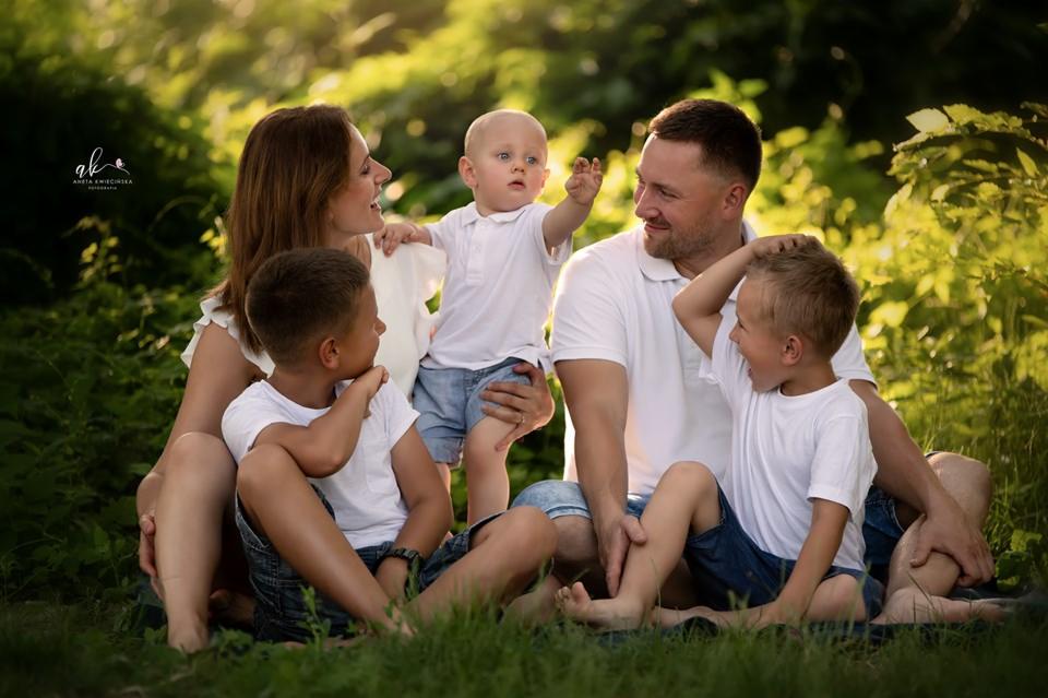 Rodzina: mama, tata i trzech synów w białych koszulkach i dżinsowych szortach, siedzą na trawie w parku, mały chłopiec wyciąga rączkę.