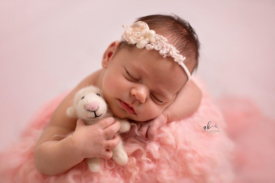 Śpiąca noworodka z kwiatową opaską na głowie, przytulająca pluszową owieczkę na różowym futerkowym tle.