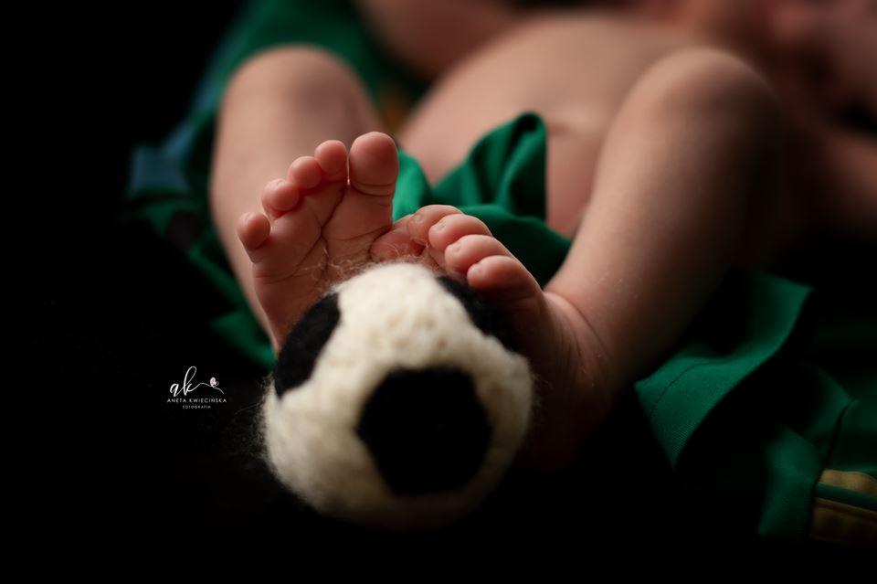 Nóżki niemowlęcia dotykają wełnianej piłki nożnej na tle zielonego materiału, sesja noworodkowa.