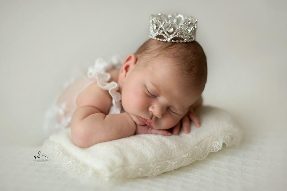 Śpiąca noworodek w koronie na poduszce z koronką, ubrana w białą sukienkę, delikatne oświetlenie, jasne tło.