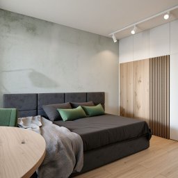 Ack Projekt - Nowoczesna sypialnia z szarą tapicerowaną ramą łóżka, zielonymi poduszkami i drewnianą podłogą. Minimalistyczne oświetlenie punktowe i szafa z drewnianymi akcentami.