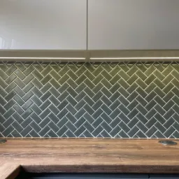 Ciemnozielone płytki ułożone w jodełkę na ścianie nad drewnianym blatem, oświetlone listwą LED pod szafkami.