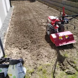 Czerwony glebogryzarek spalinowy przygotowuje glebę pod trawnik, widoczne dwa worki ziemi, obok ogrodzenie z prefabrykatów betonowych, w tle biała ściana budynku.