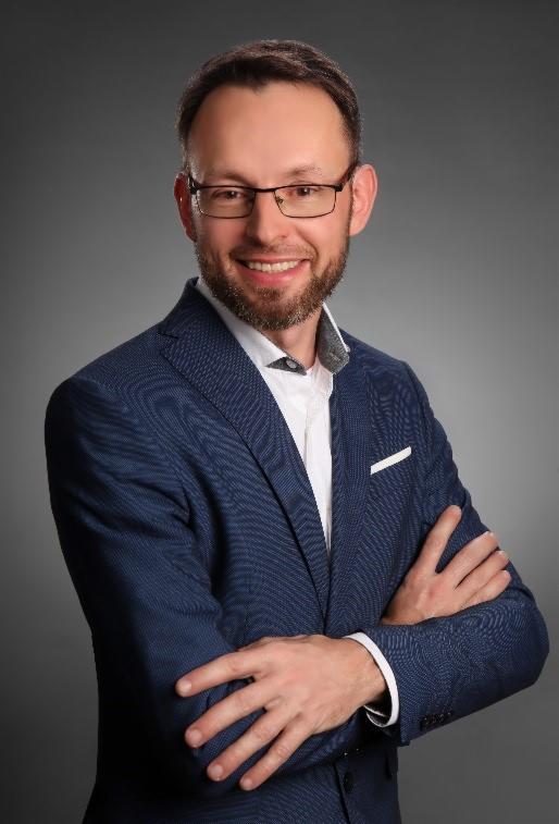 Uśmiechnięty mężczyzna w okularach i niebieskiej marynarce na szarym tle, z rękami skrzyżowanymi, prezentuje profesjonalny wizerunek.