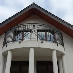 Elewacja budynku z balkonem o półokrągłej konstrukcji z kolumnami i czarną, kutą balustradą, widok z dołu na tle pochmurnego nieba.