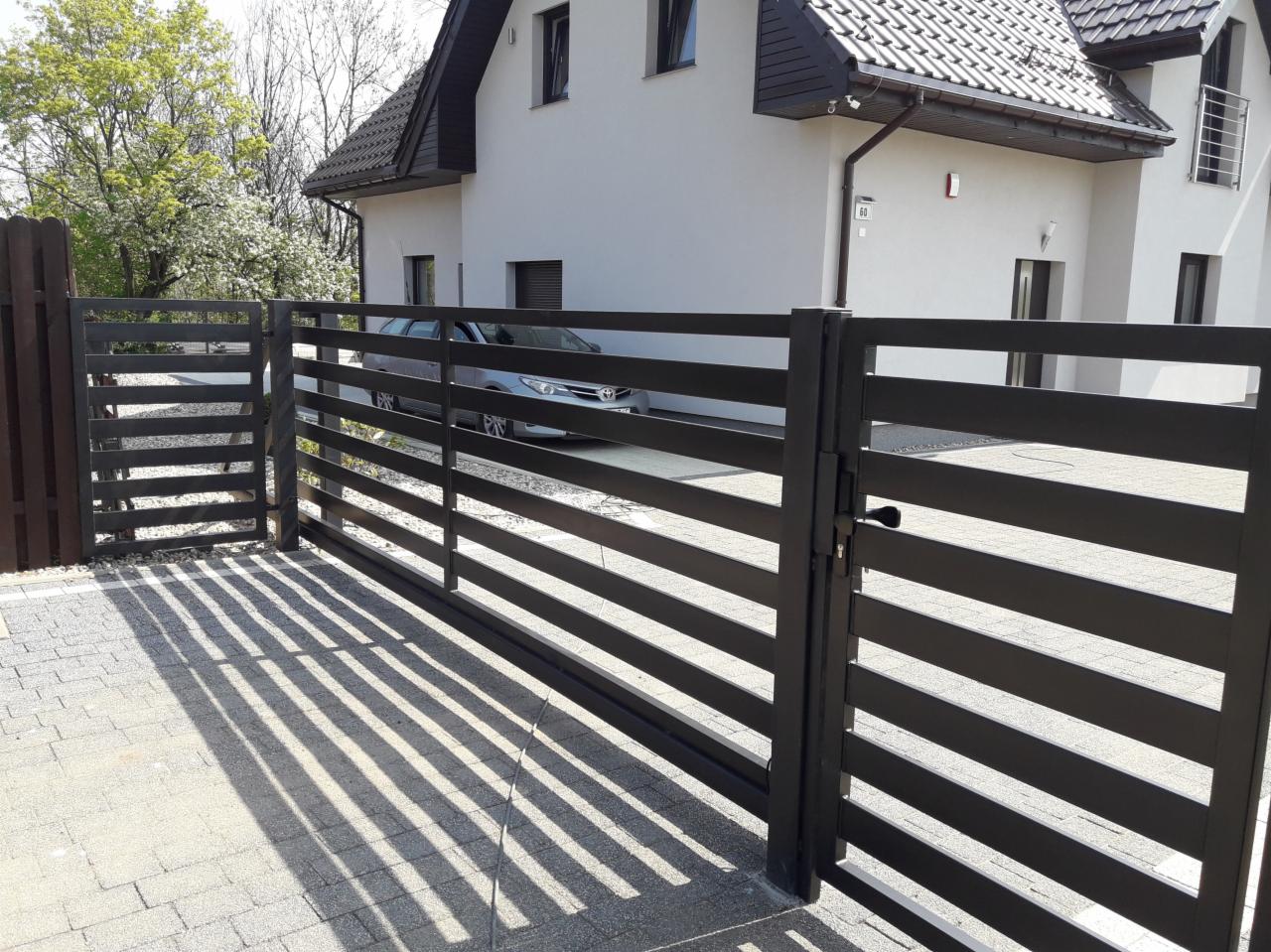 Nowoczesna, pozioma brama wjazdowa z metalu w kolorze antracytowym, widok z zewnątrz, na podjeździe widoczne cienie rzucane przez konstrukcję, w tle biały dom z szarą dachówką i zaparkowany samochód.